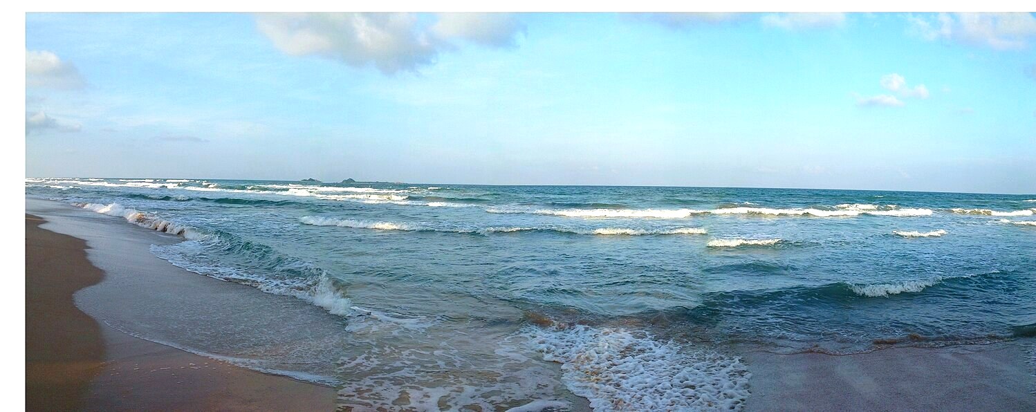 Panoramic view of waves washing onto Nilaveli Beach beneath a blue, cloud-dotted sky.